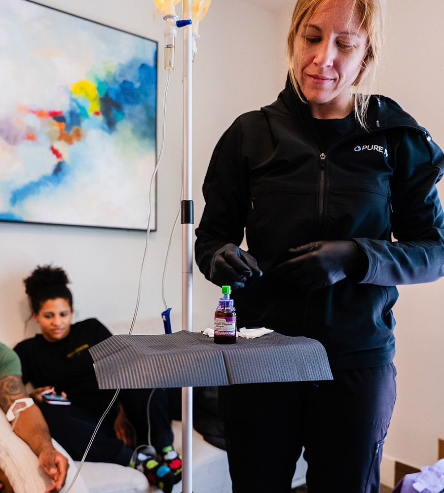 A person in a black jacket and gloves prepares an IV medication vial next to a patient receiving treatment on a sofa.