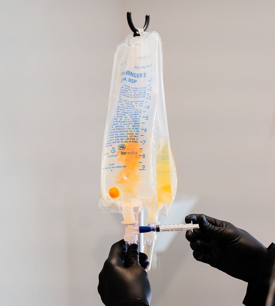 IV bag with yellow liquid, hanging from a hook. Gloved hands holding a syringe inject medication into the bag.