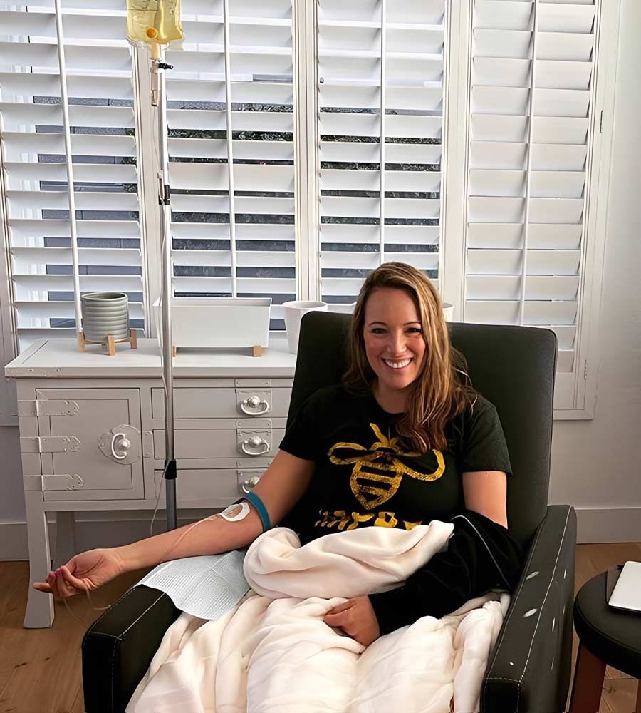 Woman receiving IV treatment for headache, smiling, in a chair. Arm has bandage and tourniquet. White room with shutters.