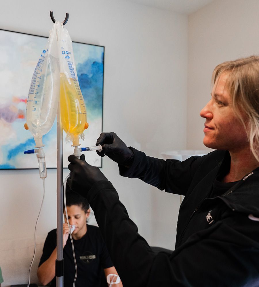 Woman in black gloves adjusts IV bag hanging from a stand; a person sits nearby, receiving treatment.
