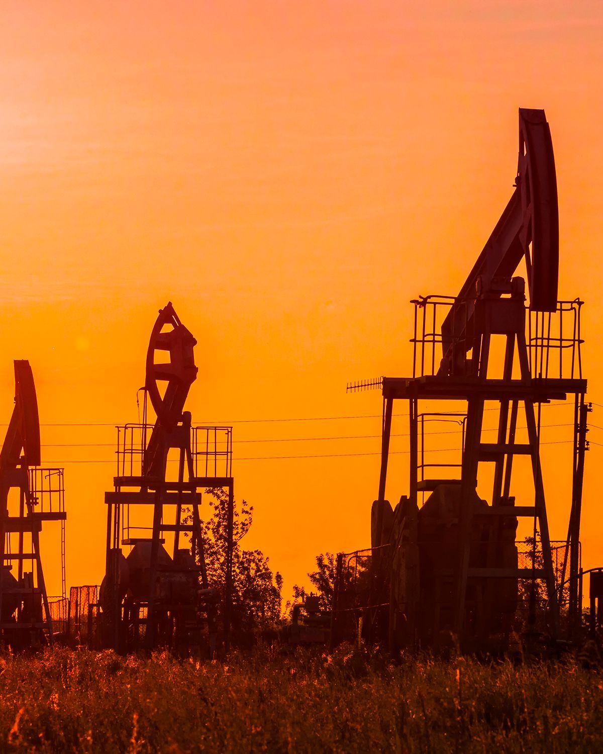 Silhouettes of several oil pumpjacks standing in a field against a bright, orange sunset sky.