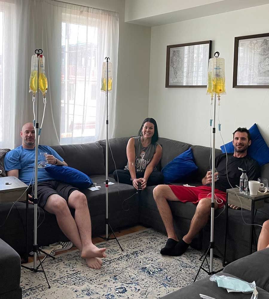 Three people receiving IV infusions on a couch, medical equipment in a living room setting.