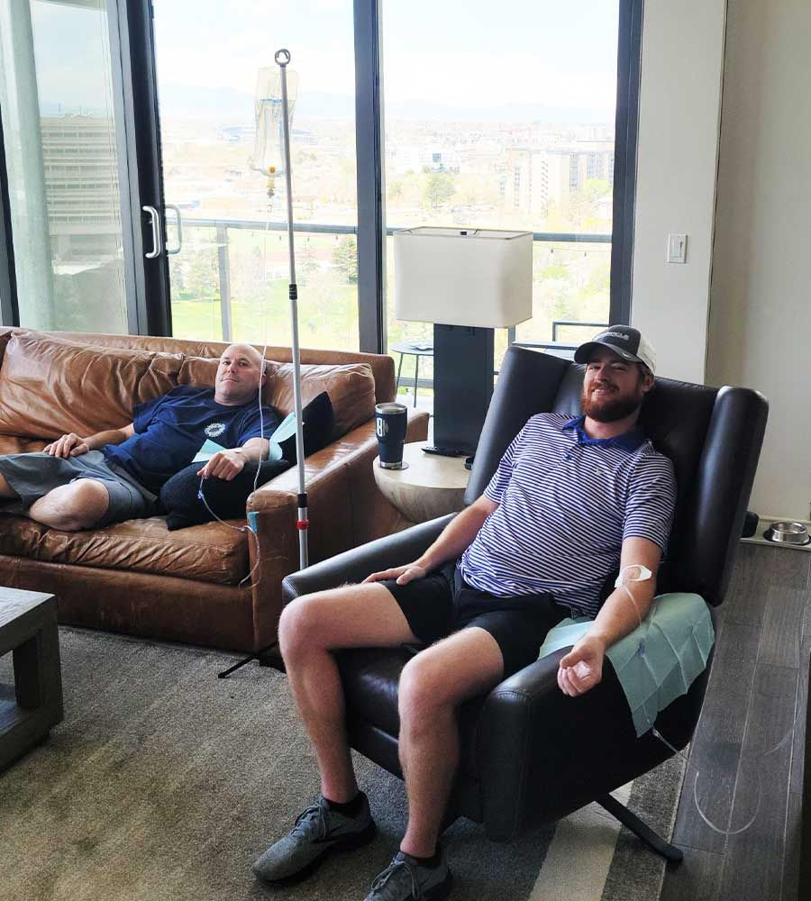 Two men receiving IV infusions in a modern living room. One sits in a chair, the other reclines on a leather sofa.