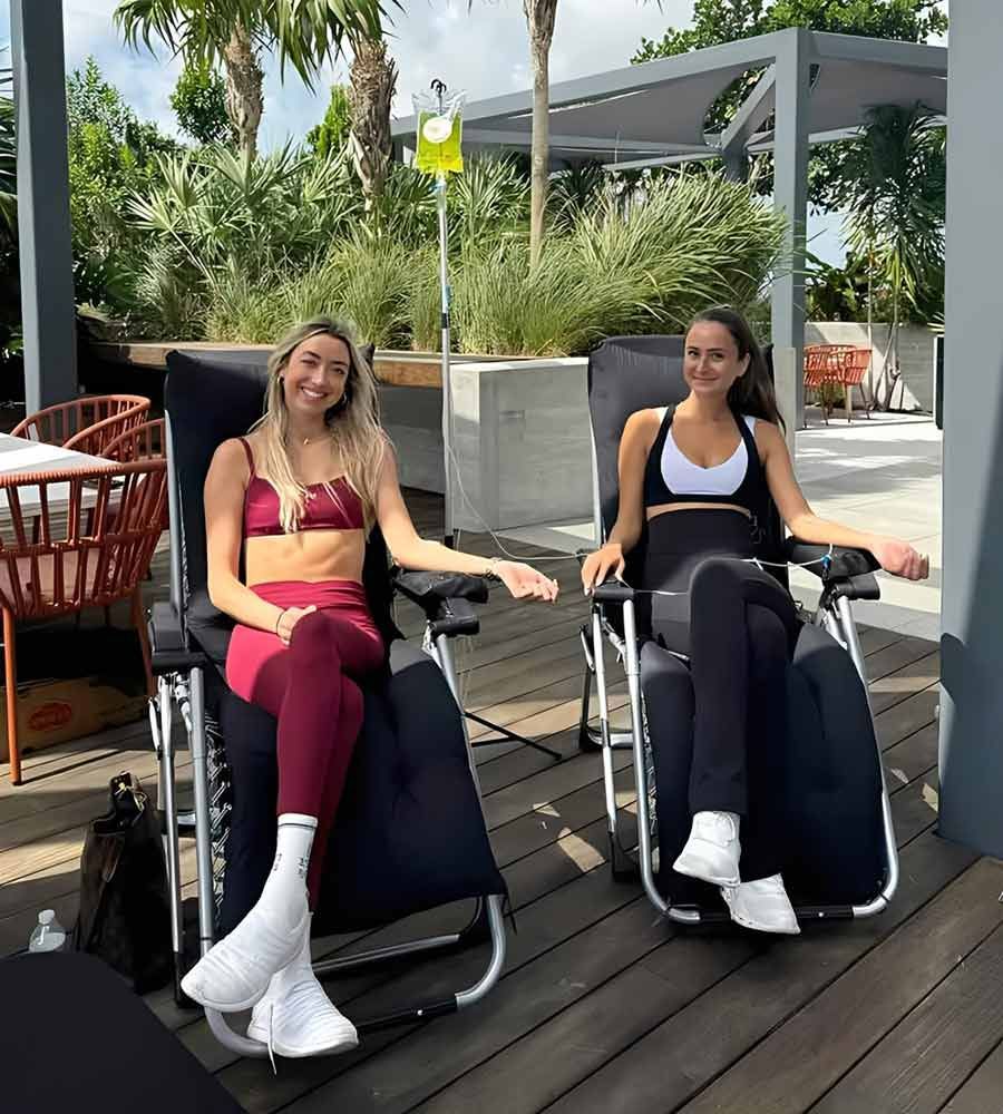 Two women in fitness attire receiving IV hydration therapy outdoors after a workout.