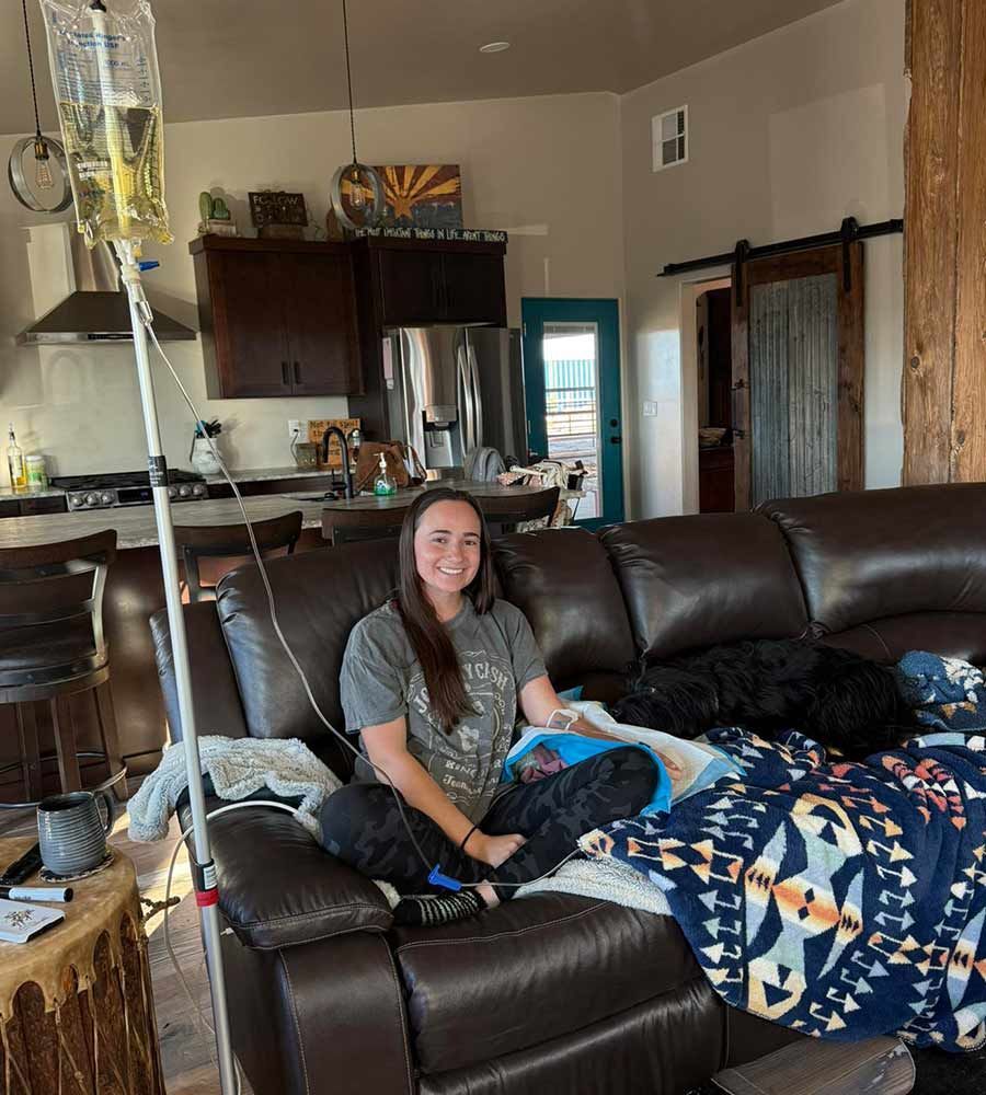 Woman on couch with IV drip in home setting. Smiling, with dog, blanket, and kitchen visible.