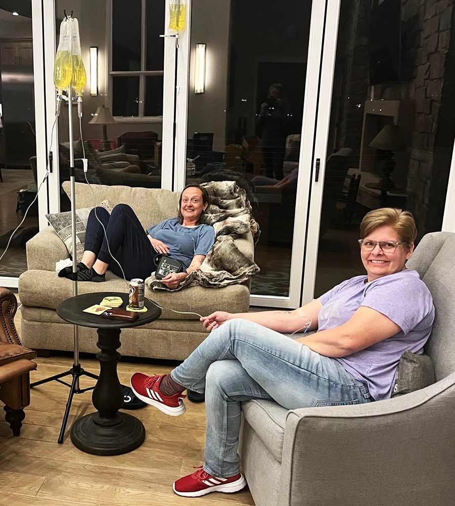 Two people relax in armchairs, receiving IV infusions in a modern living room setting.