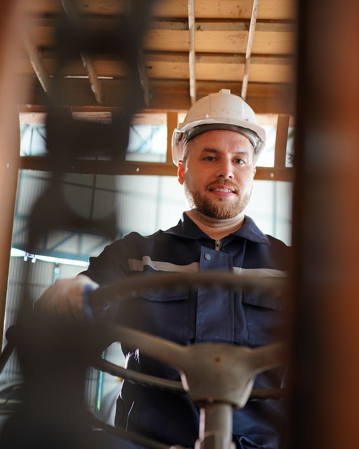 A worker in a hard hat and blue uniform sits in the driver's seat of a forklift, looking forward.