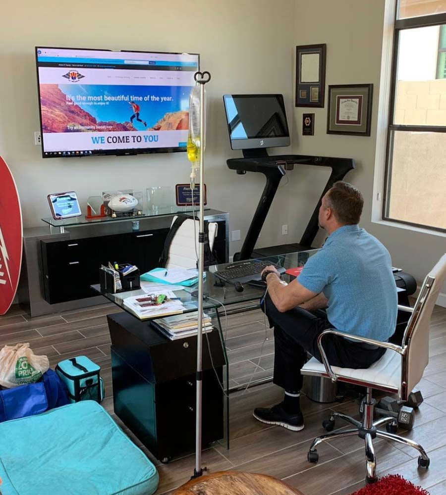 Man working at desk with an IV drip. Office has a computer, monitor, and treadmill.