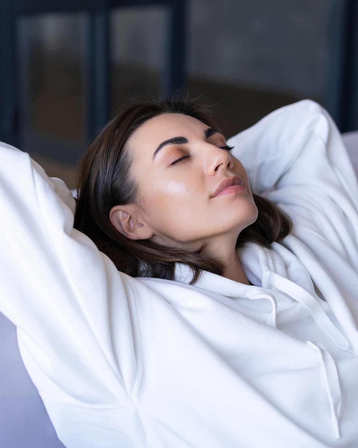 Person with dark hair in a white hoodie relaxing with hands behind head and eyes closed.