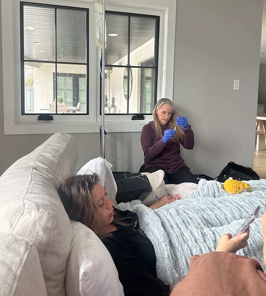 Person receiving IV fluids on couch, another person preparing something, near a window.