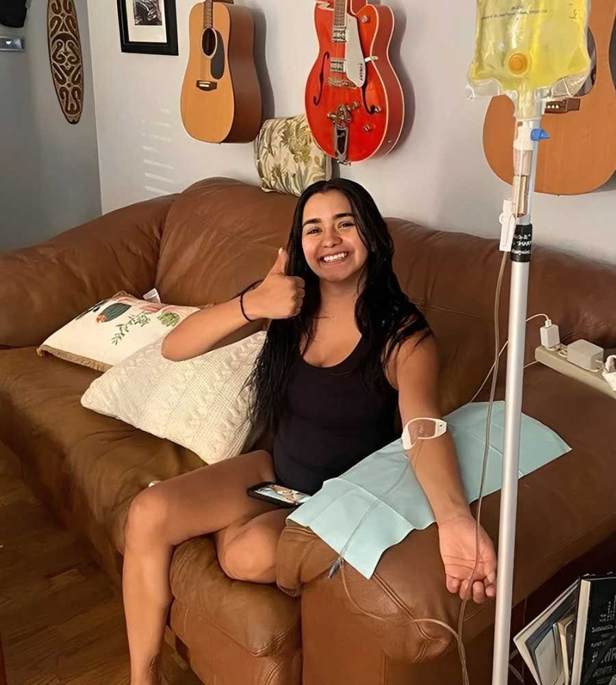 Woman on couch with IV drip, giving thumbs up. Guitars on wall.