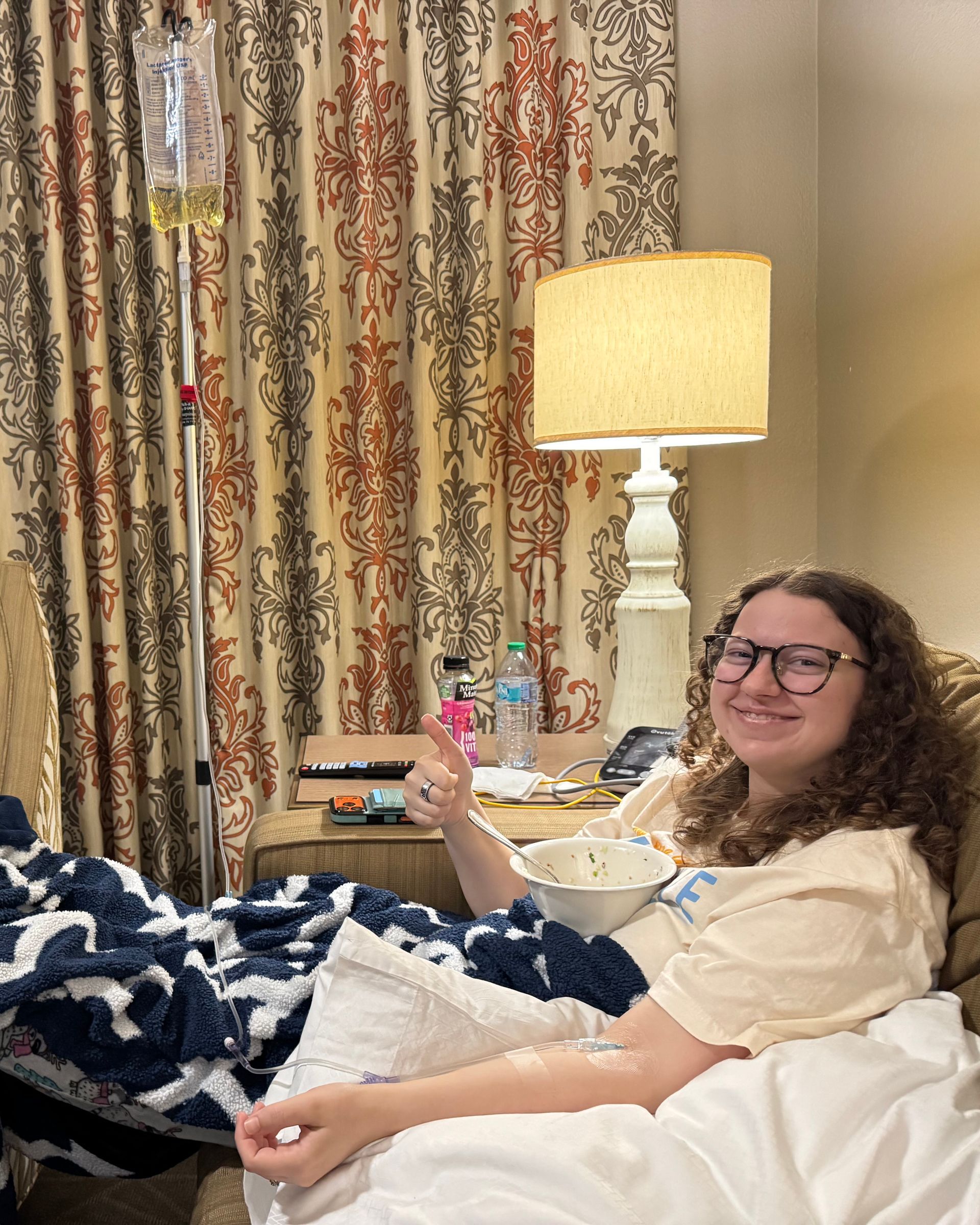 Person with curly hair and glasses smiling, giving thumbs up while receiving IV fluids indoors