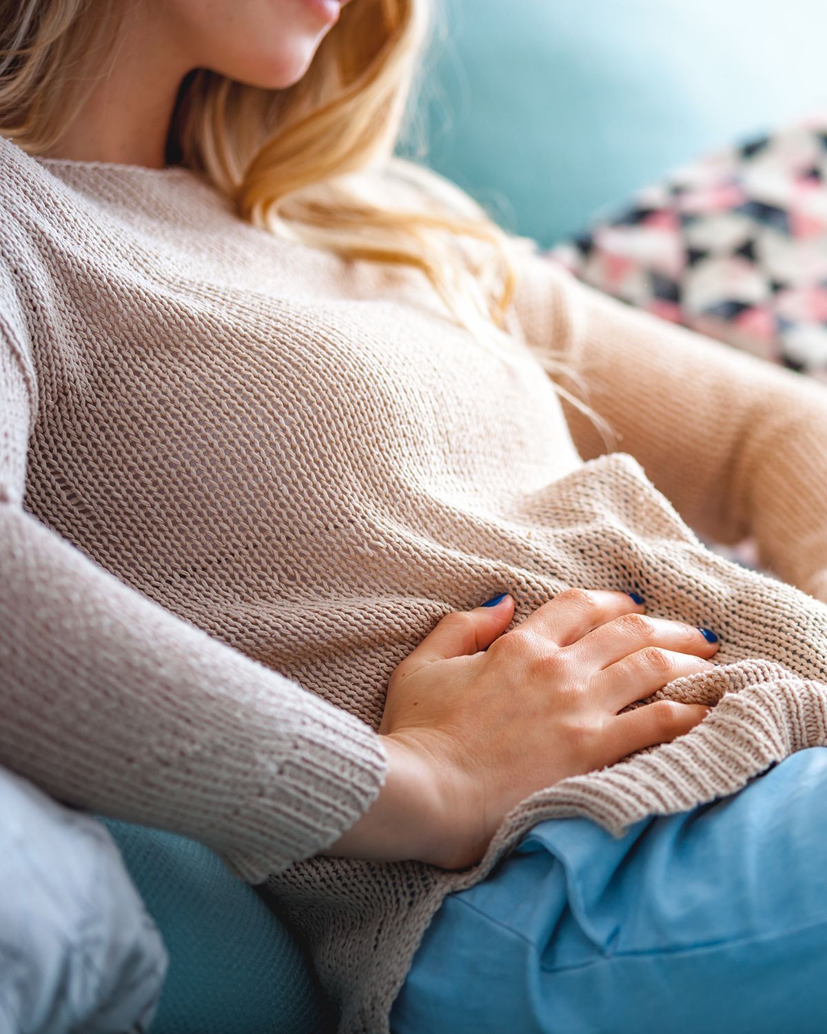 A person wearing a beige sweater resting their hand on their abdomen while sitting on a couch.