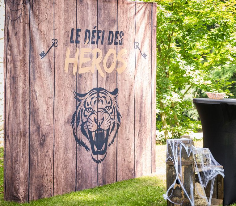 Fond en bois avec inscription « Le Défi des Héros », tête de tigre et clés. Herbe verte, arbres et table visibles.
