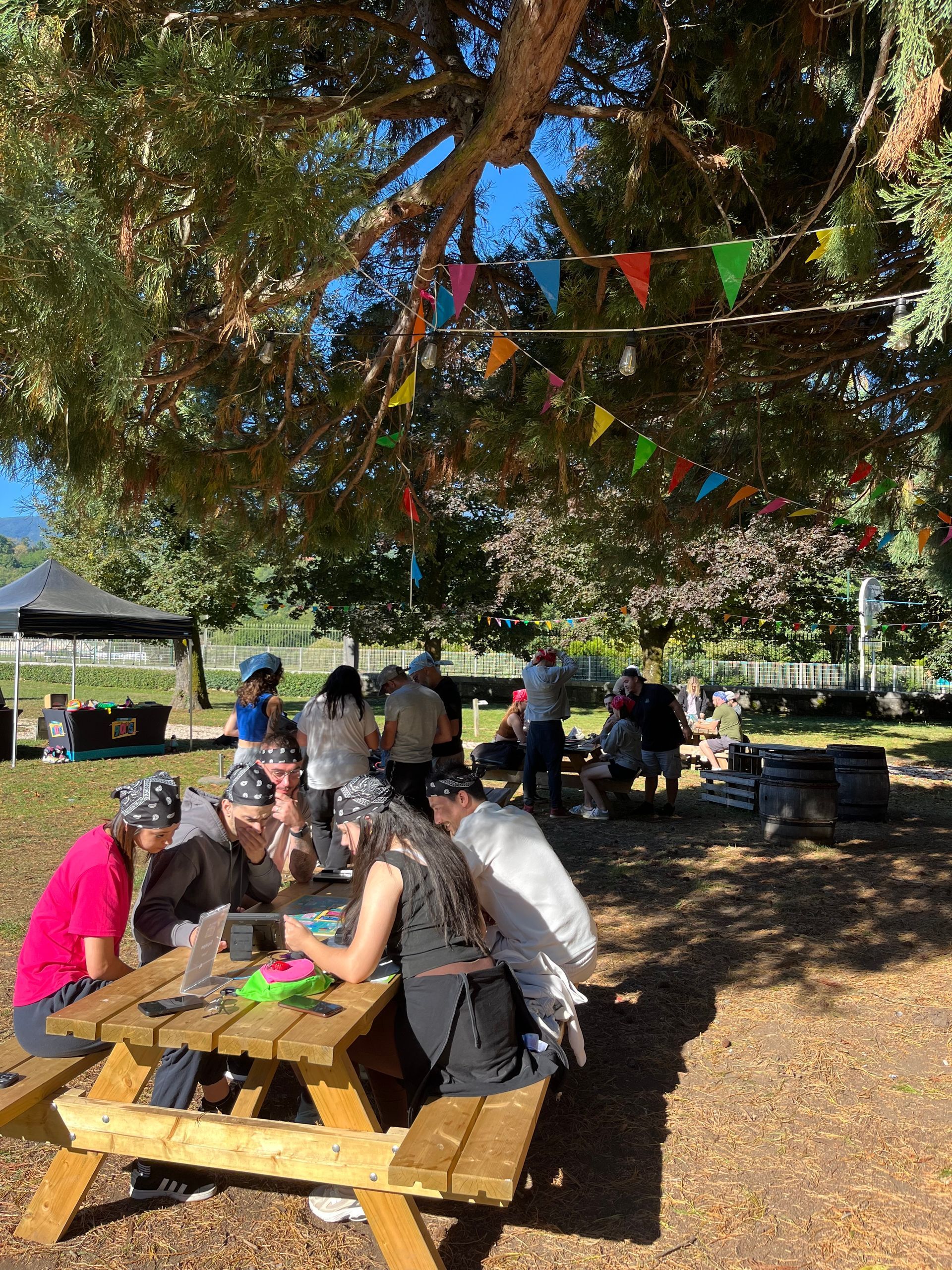 Les gens se sont rassemblés autour de tables de pique-nique sous un arbre avec des drapeaux colorés au-dessus.