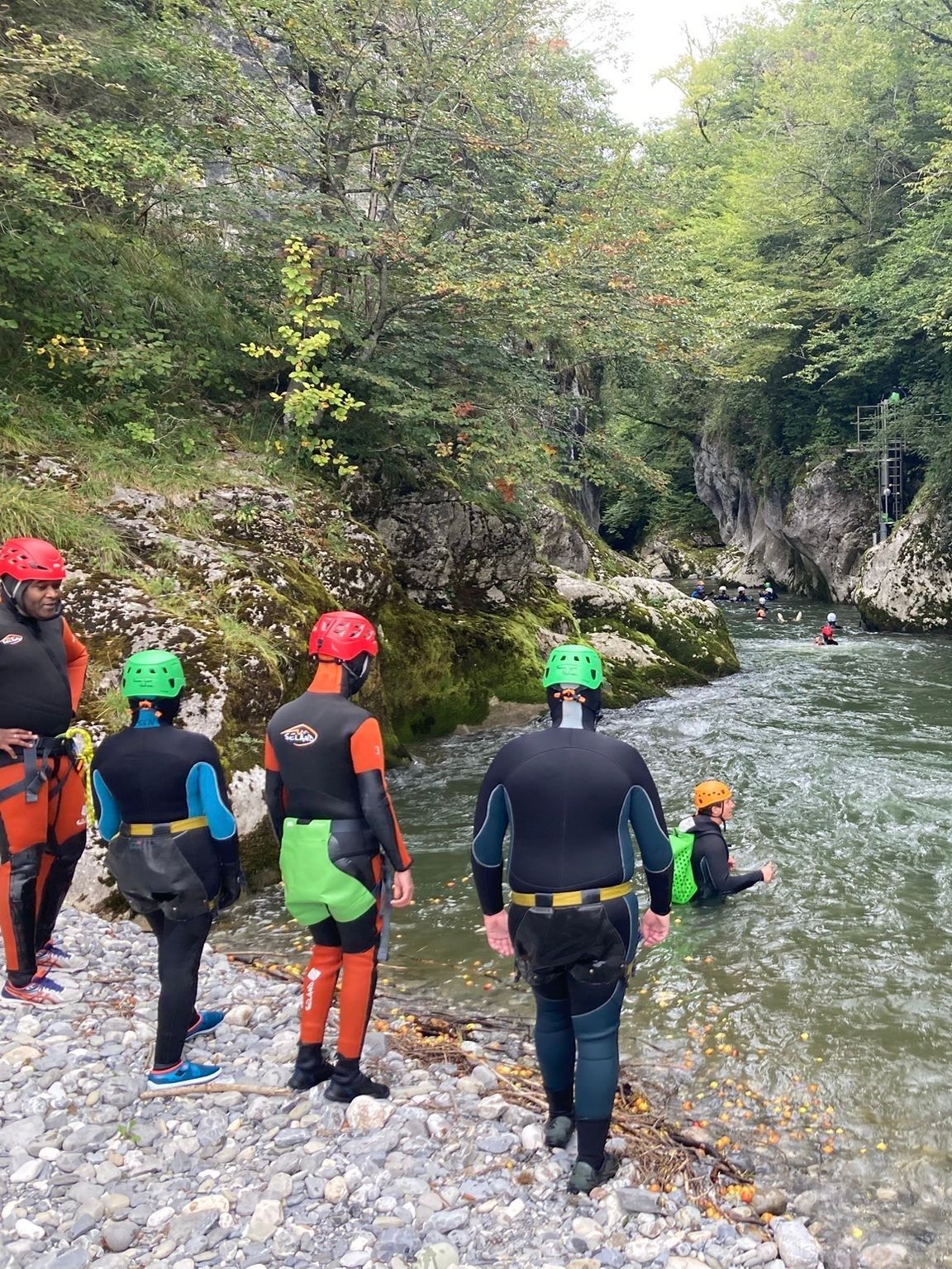 Des gens en combinaison de plongée près d'une rivière ; une personne est dans l'eau. Rivage rocheux avec des arbres.