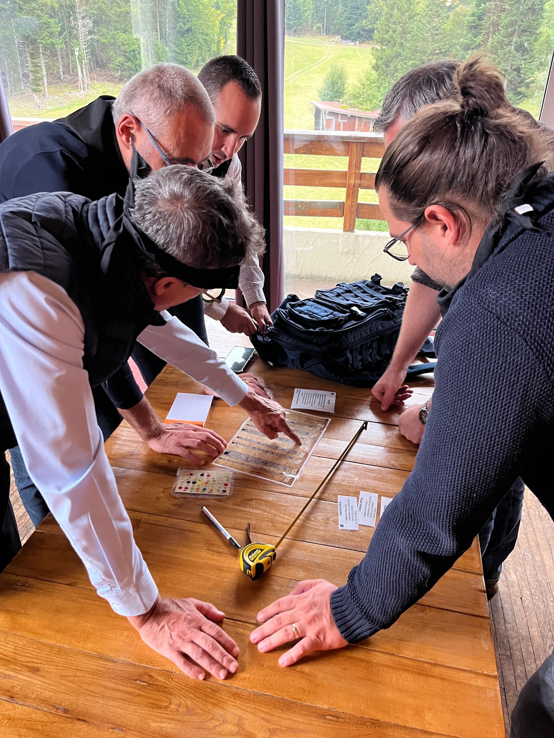 Des personnes autour d'une table, l'une d'elles ayant les yeux bandés, pointent des objets. À l'intérieur, table en bois.