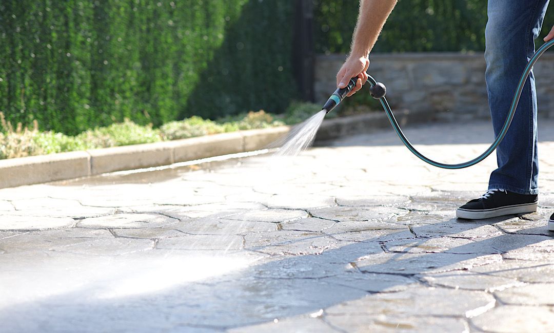 Person spraying water from a hose onto a sunlit paved driveway