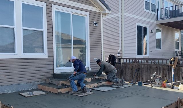 Two workers install pavers beside a house with a sliding glass door and porch steps.