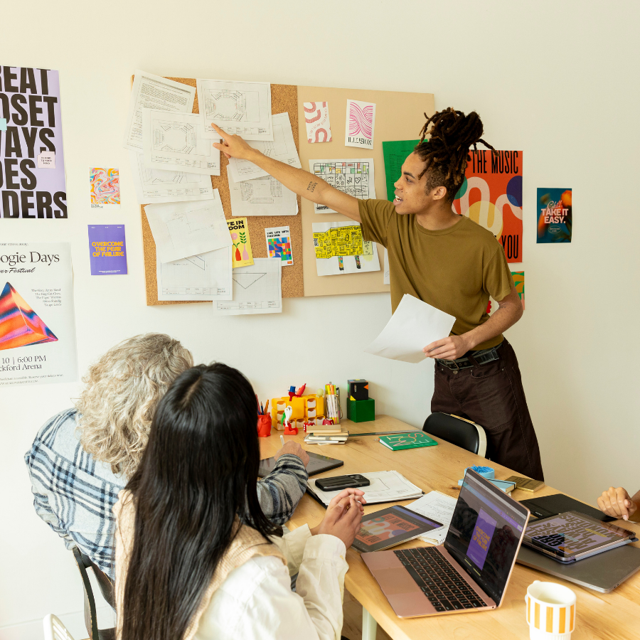 Person points at a wall of notes during a team brainstorming session at a desk with laptops and papers.
