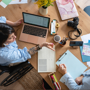 Two people collaborating at a wooden desk with laptops, notebooks, camera, and coffee in a bright workspace