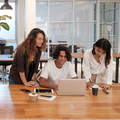 Three coworkers smiling around a laptop at a wooden table in a bright office space