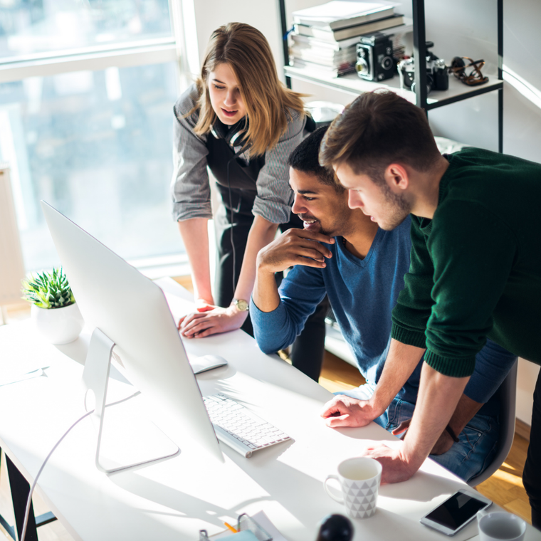 Three coworkers lean over a desktop computer in a bright office, collaborating on a project.