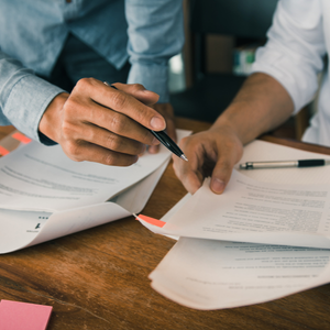 Two people review paperwork at a wooden table, pointing and writing with pens.