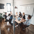 Team meeting around a table in a bright office, with laptops, notes, and a whiteboard in the background.