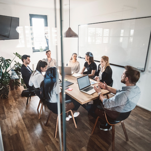 Team meeting around a table in a bright office, with laptops, notes, and a whiteboard in the background.