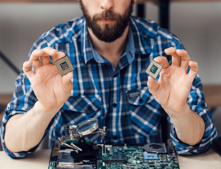 Man holding two computer processors above a circuit board, with a magnifying glass.