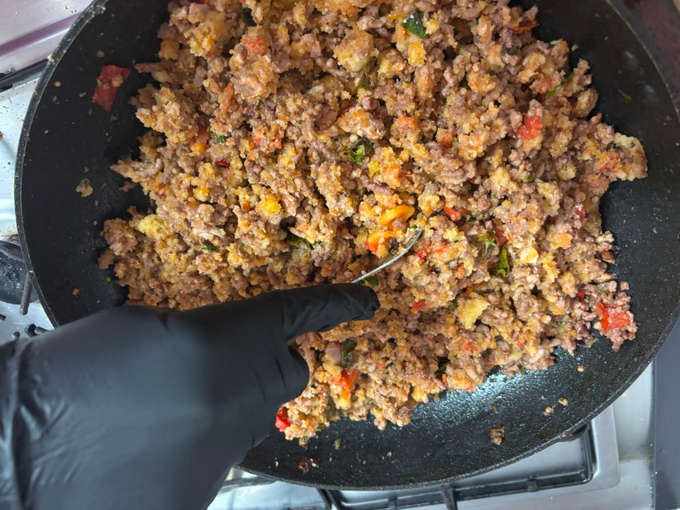 Mano guantata che mescola carne macinata e verdure rosolate in una padella sul fornello