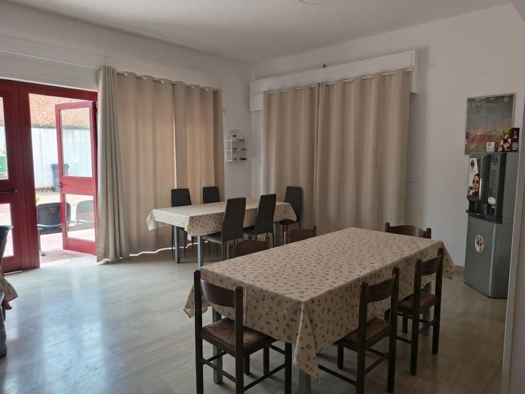 Luminosa sala da pranzo con due tavoli, sedie, tende beige e una porta a vetri che dà su un balcone rosso.