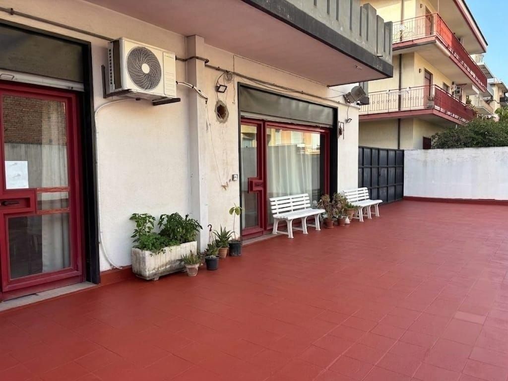 Patio dell'appartamento con pavimento in piastrelle rosse, sedie bianche, piante in vaso e porte e finestre con finiture rosse.