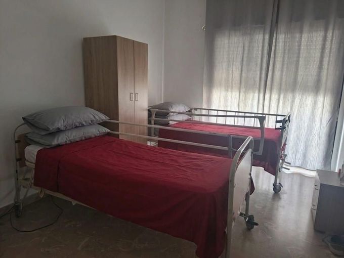 Stanza d'ospedale con due letti, coperte rosse, tende grigie e un armadio.