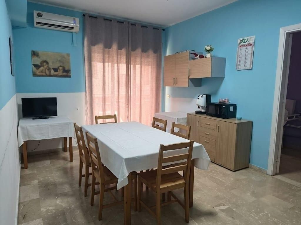 Piccola sala da pranzo con pareti blu, tavolo, sedie, TV e condizionatore, illuminata da una finestra con tenda.