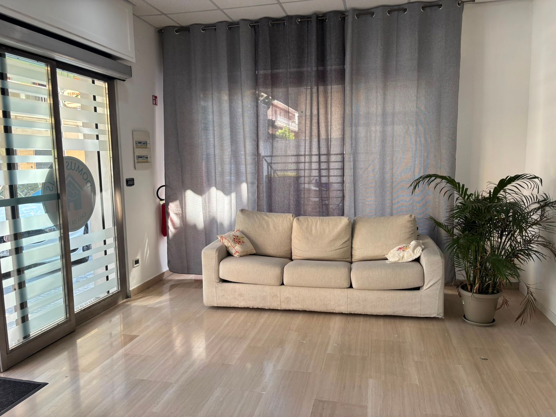 Luminoso soggiorno con divano beige, tende grigie, porta scorrevole in vetro e pianta in vaso su pavimento in legno.