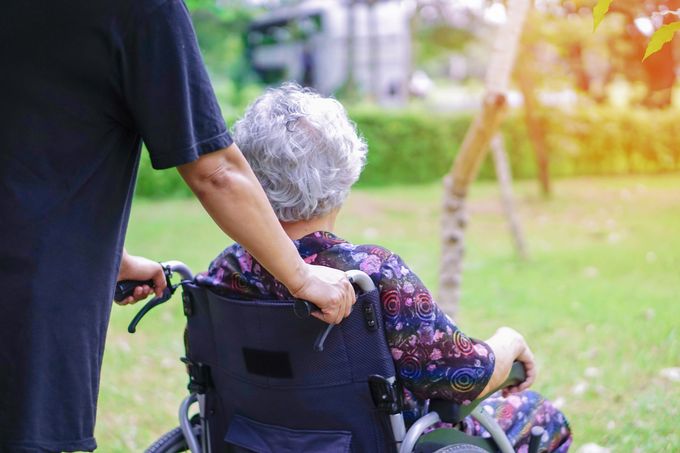 In un parco soleggiato, una persona spinge una sedia a rotelle, con a bordo un passeggero anziano che indossa una camicia viola a fantasia.