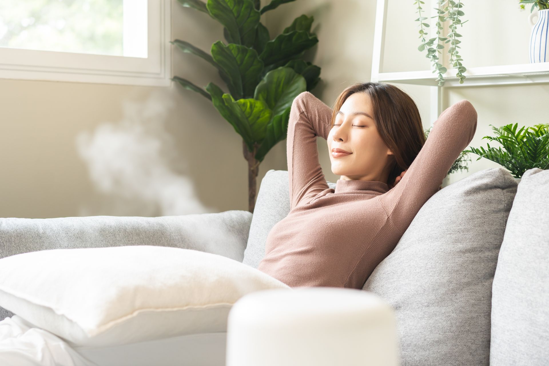 A person relaxes on a sofa with hands behind their head, near a steaming humidifier in a bright, plant-filled room.