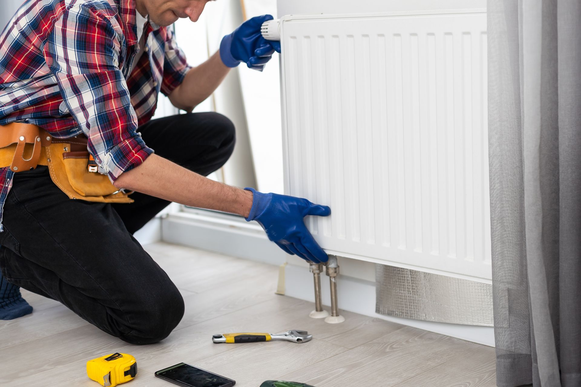 A technician in a plaid shirt and work belt kneels on the floor, installing a white radiator onto wall pipes.