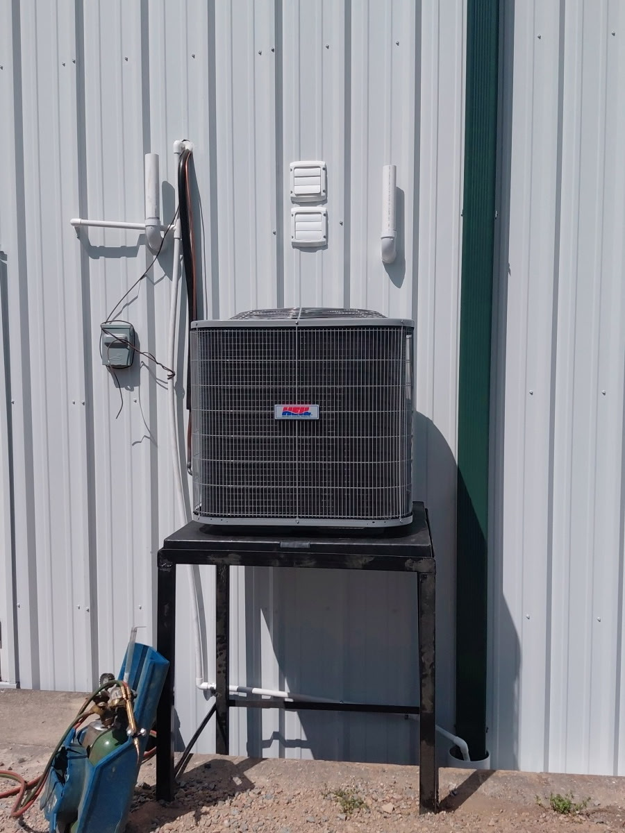 An outdoor HVAC unit sitting on a black metal stand against a white corrugated metal wall.