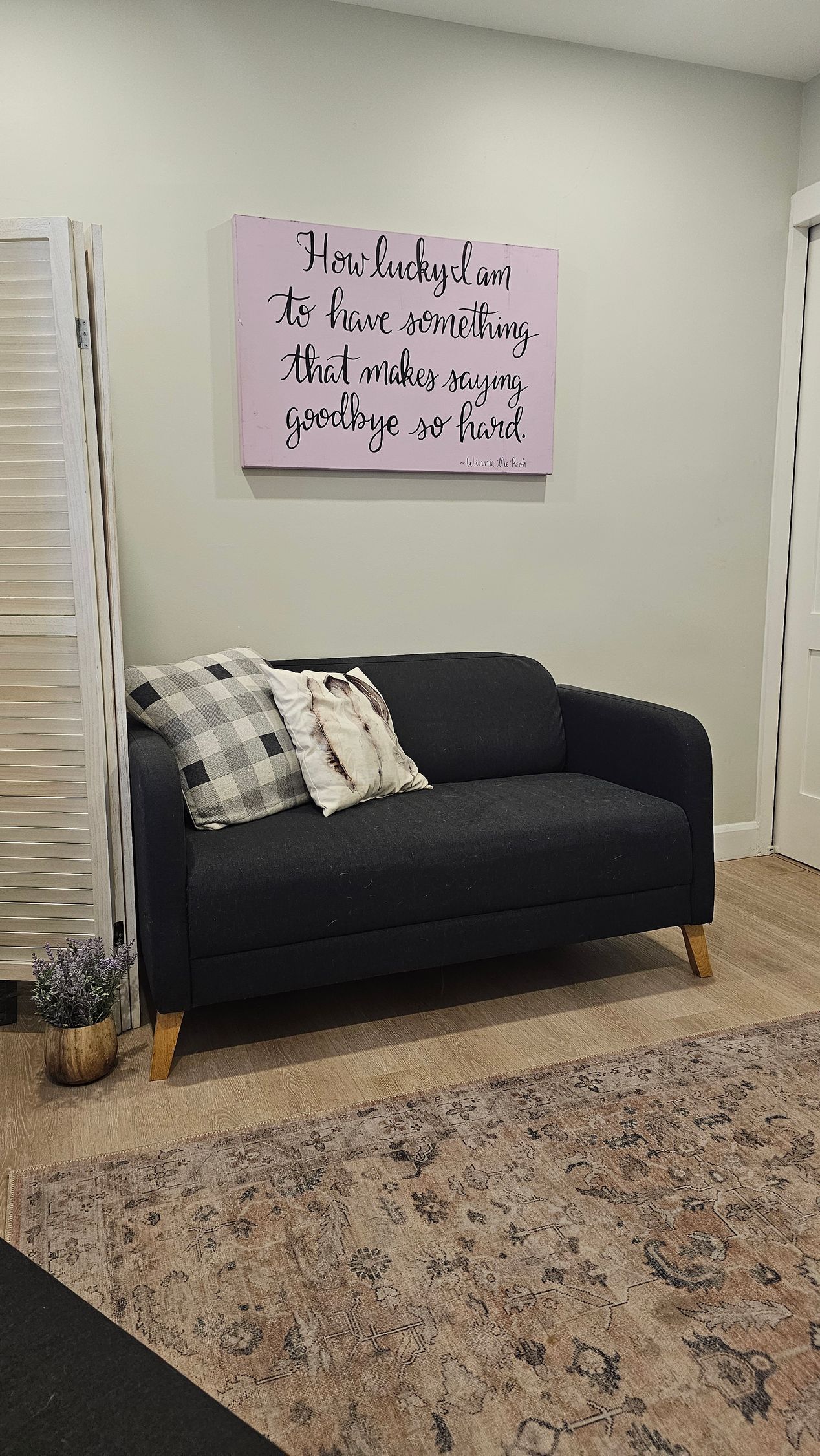 Black loveseat with pillows, beneath a framed pink sign. Beige and brown rug.