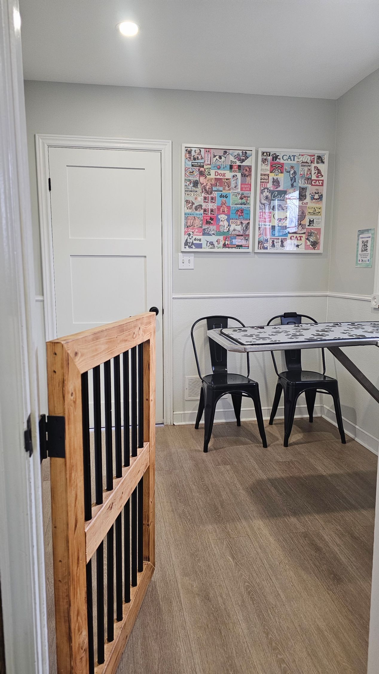 Wooden dog gate in doorway, leading to a small dining area with a table, chairs, and two framed collages on the wall.