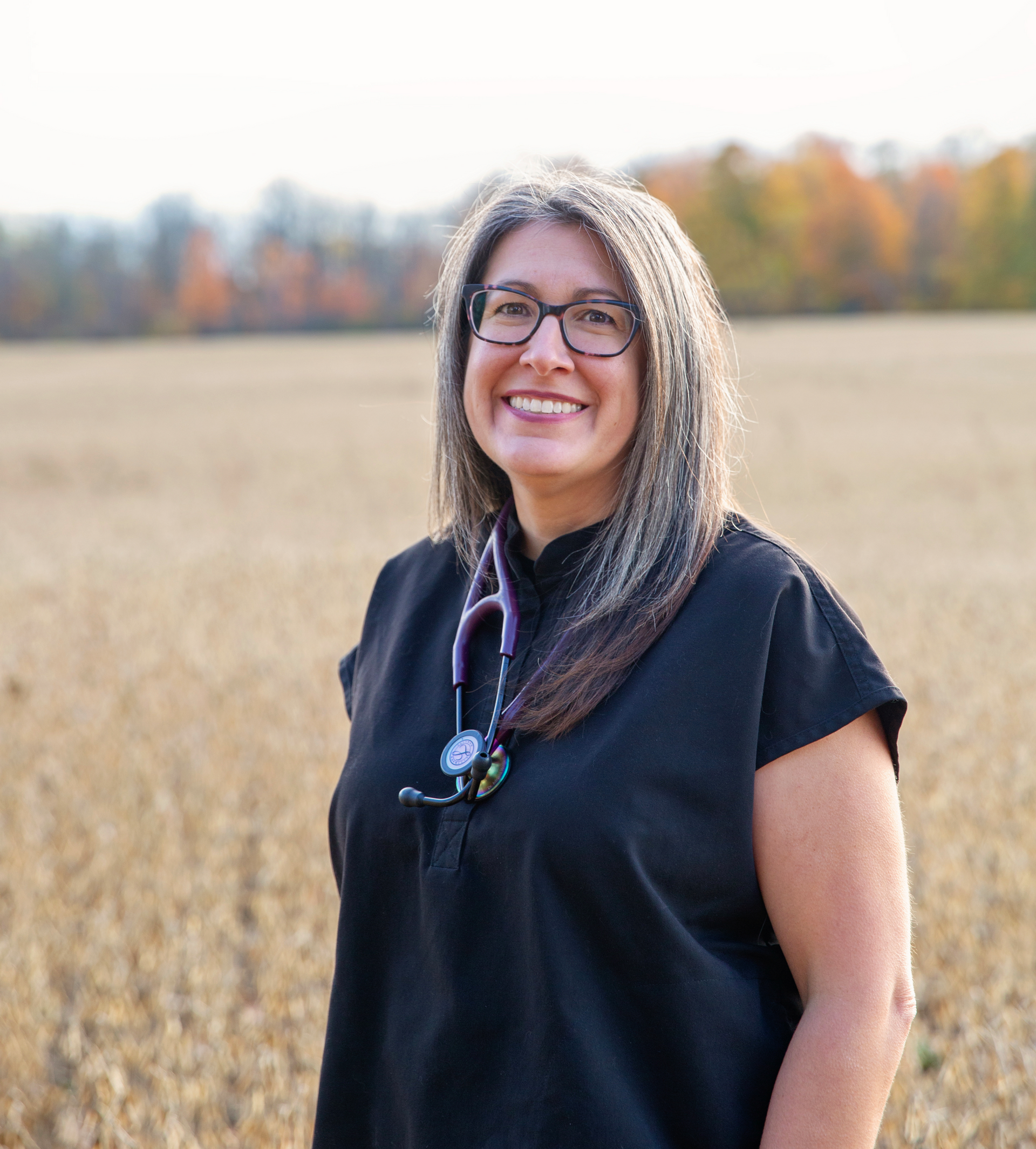 Woman with glasses and stethoscope smiles in a field.