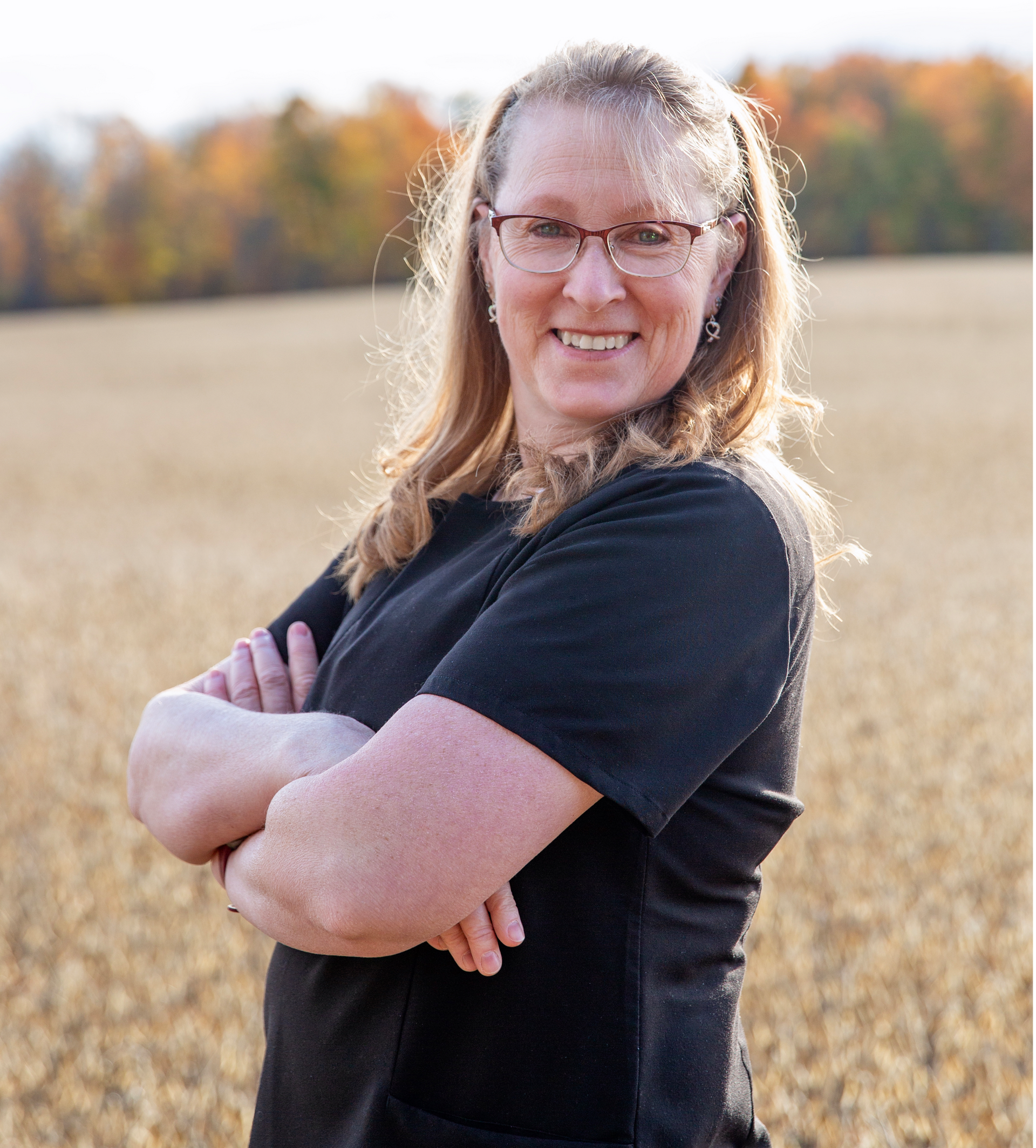 Woman in glasses with arms crossed, smiling in a field with a blurred autumn background.