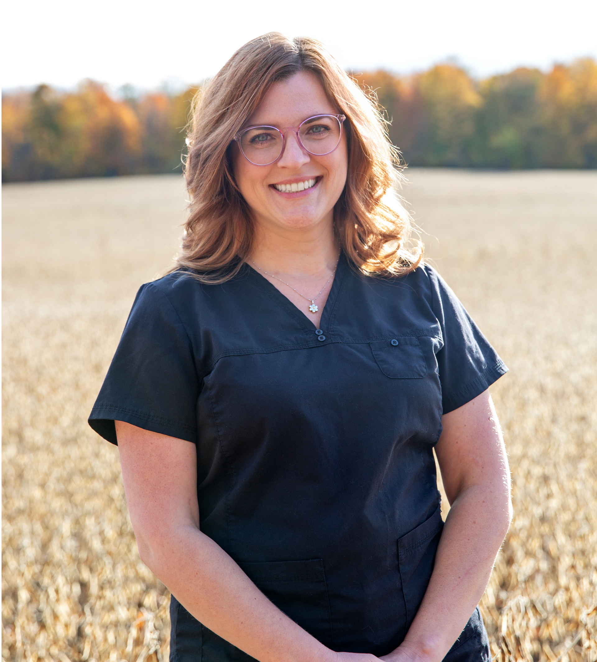 Smiling woman in black scrubs, glasses, standing in a field of dry vegetation.