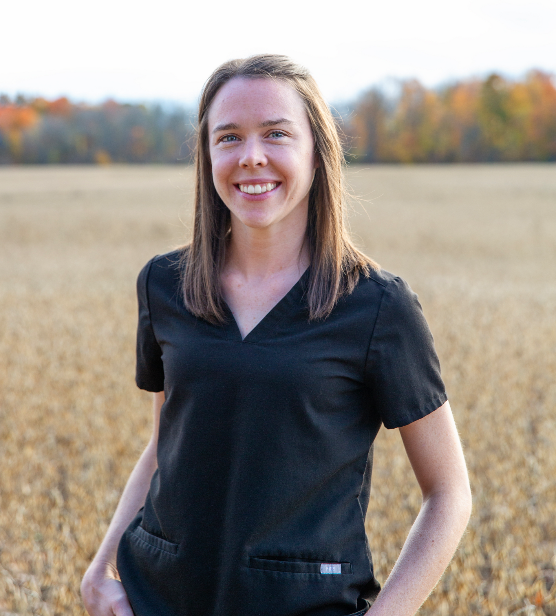 Woman in black scrubs smiling in a field; fall foliage in the background.