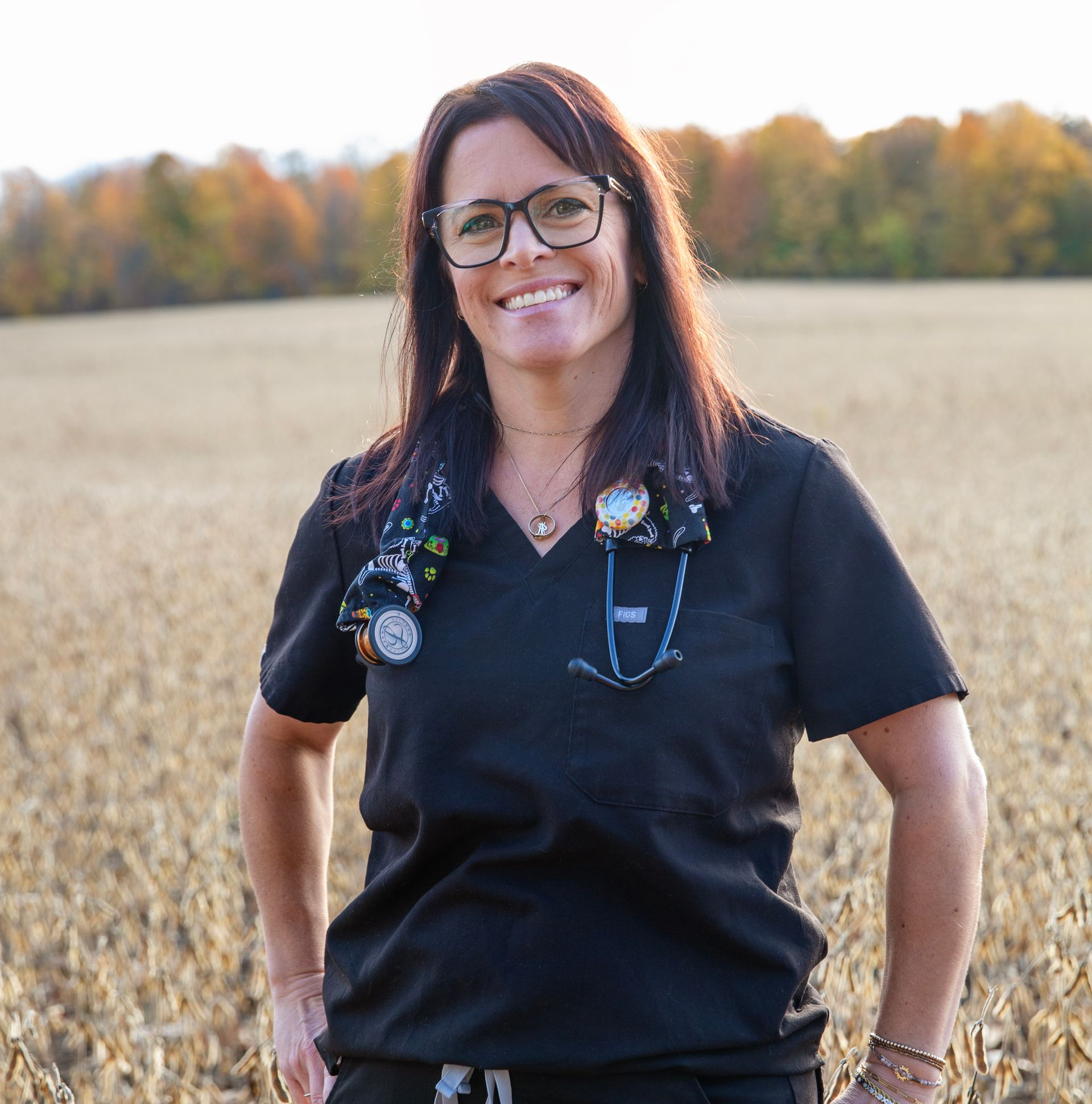 Woman in black scrubs, glasses, and stethoscope smiles in a field.