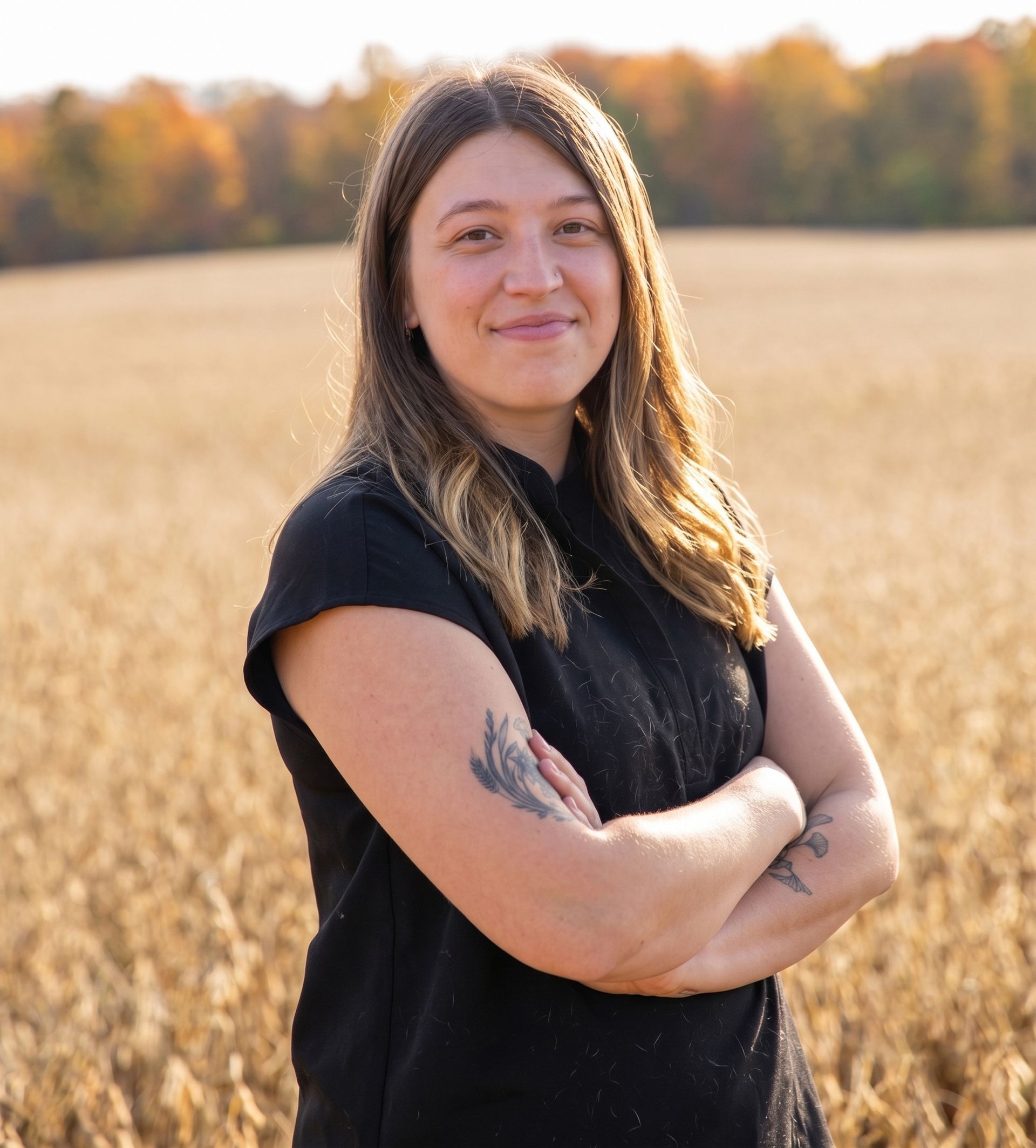 Woman with crossed arms smiles in a field, trees in the background, daytime.