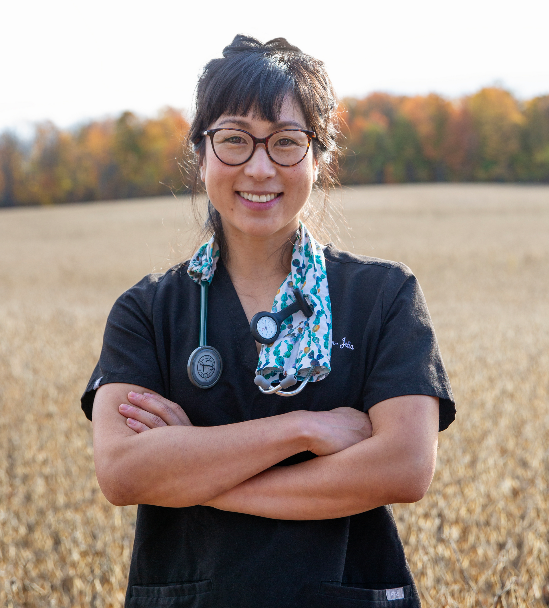 Woman in medical scrubs with stethoscope, arms crossed, smiling in a field with fall foliage.